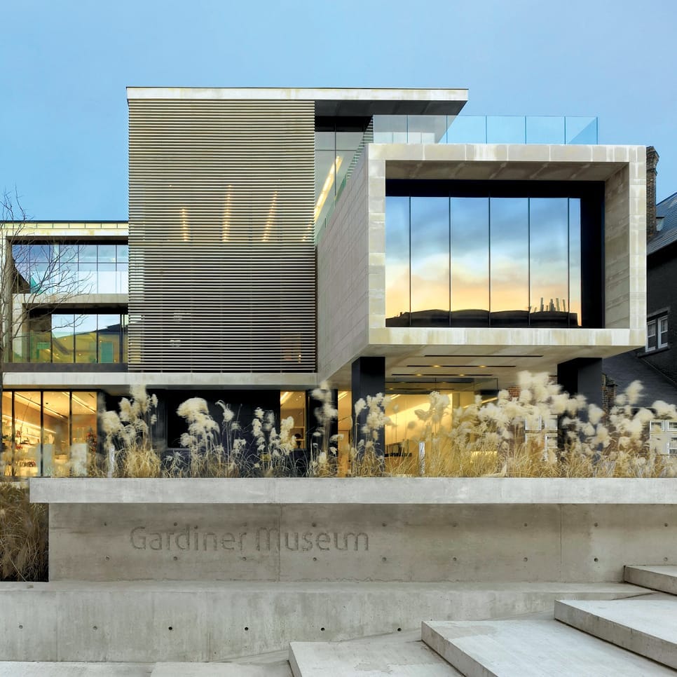 ด้านนอกที่ทันสมัยของ Gardiner Museum ด้วยสถาปัตยกรรมกระจกและหิน มีหญ้าประดับอยู่เบื้องหน้า และแสงยามเย็นอันนุ่มนวลส่องสว่างอาคาร