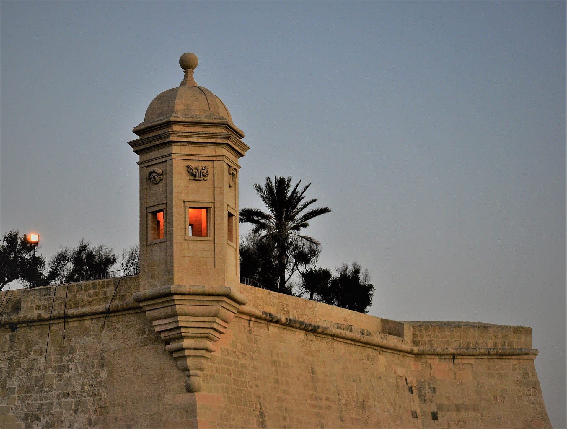 Torre di guardia in pietra nei Giardini di Gardjola a Senglea, Malta, con la calda luce della sera e palme in silhouette contro un cielo sereno.