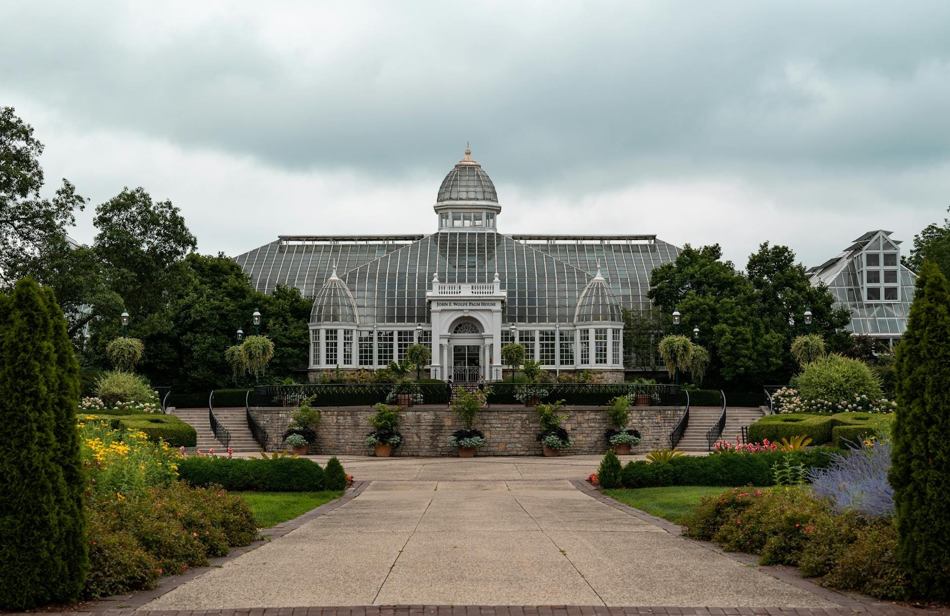 Vista ampia dell'ingresso del Garfield Park Conservatory con la cupola di vetro, incorniciata da lussureggianti giardini e un vialetto sotto un cielo drammatico e nuvoloso.