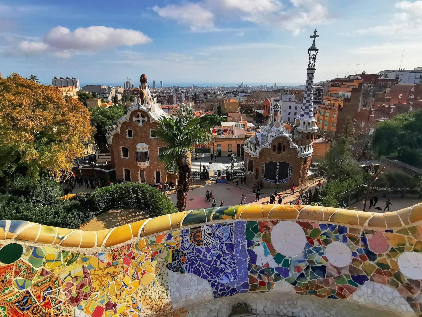 View over Park Güell’s colorful mosaic terrace, Gaudí-designed gatehouses, and the Barcelona cityscape under a blue sky.