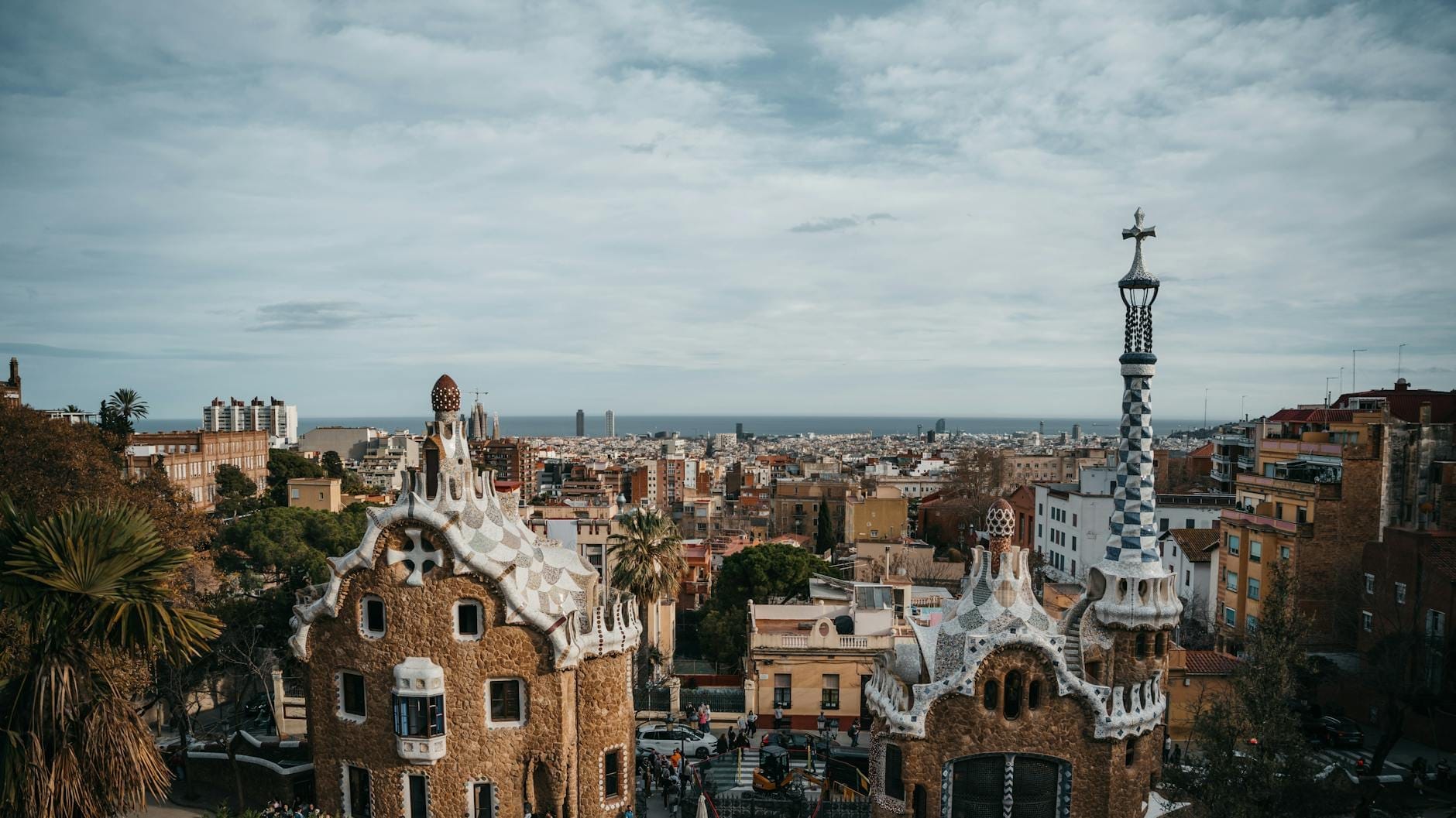 Wide-angle view over Park Güell’s colorful, whimsical houses and spires, with Barcelona cityscape and Mediterranean Sea in the background on a partly cloudy day.