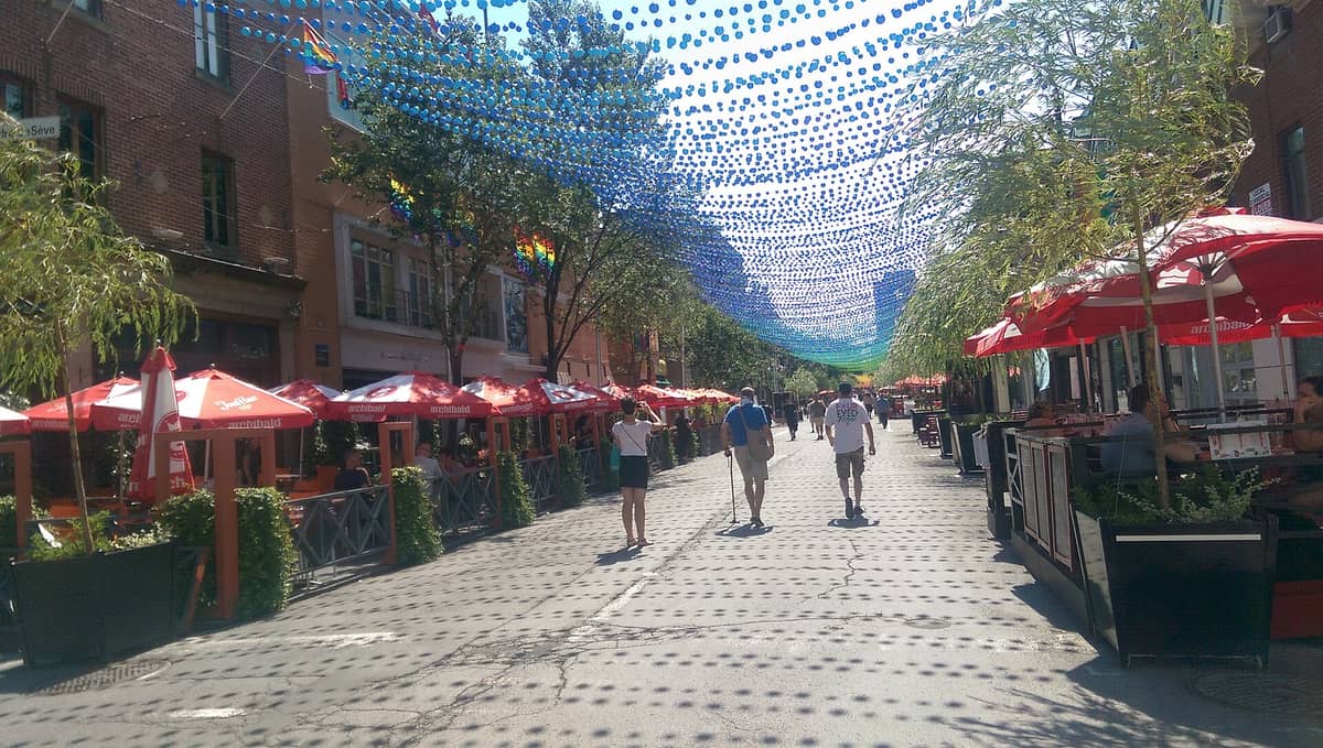 Die Fußgängerzone im Gay Village Montréal auf der Rue Sainte-Catherine mit bunten hängenden Kugeln, Außenterrassen und flanierenden Menschen.