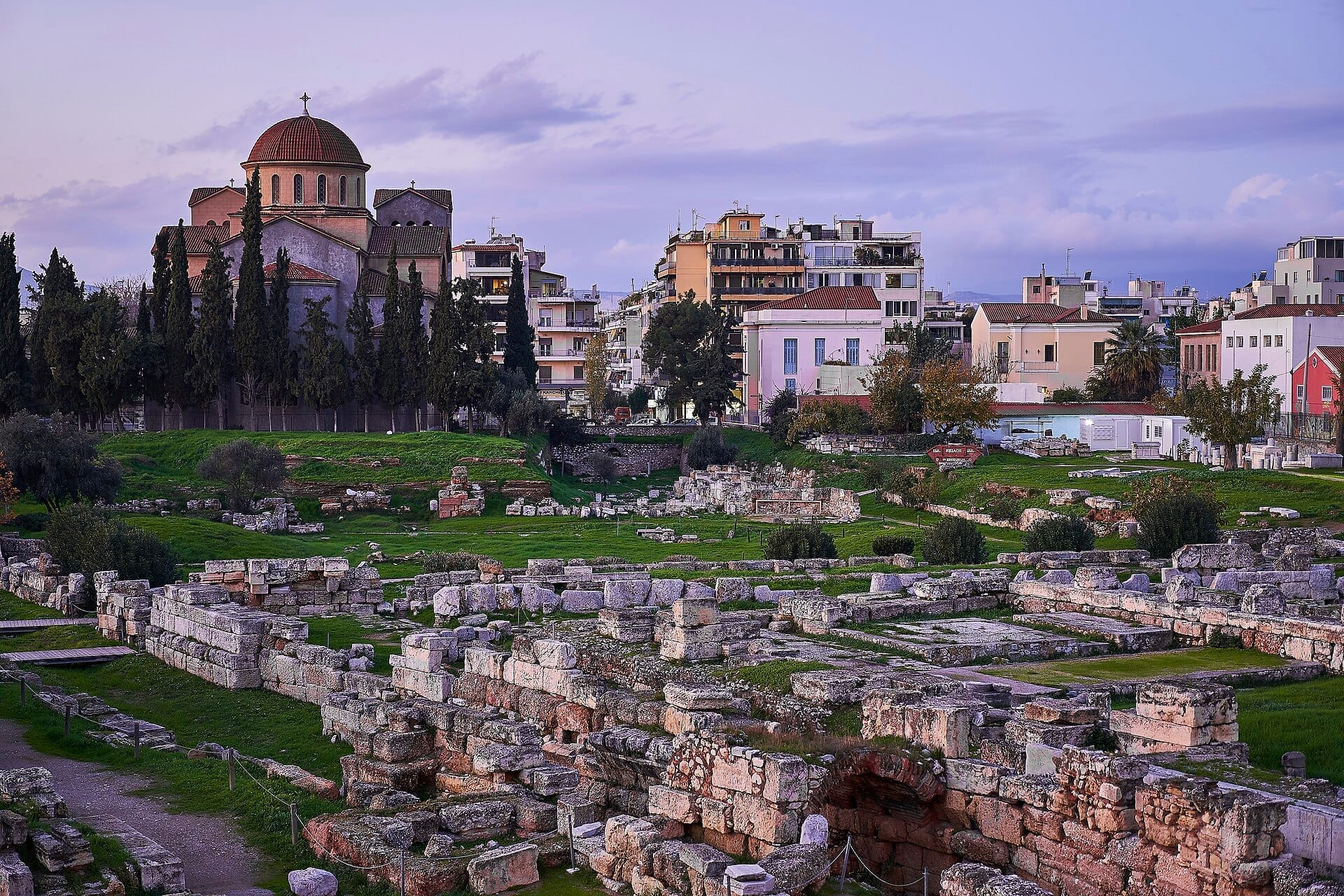 ซากปรักหักพังหินโบราณของสุสาน Kerameikos พร้อมโบสถ์หลังคาโดมและอาคารในเมืองหลากสีสันในพื้นหลังภายใต้ท้องฟ้ายามเย็นที่นุ่มนวล