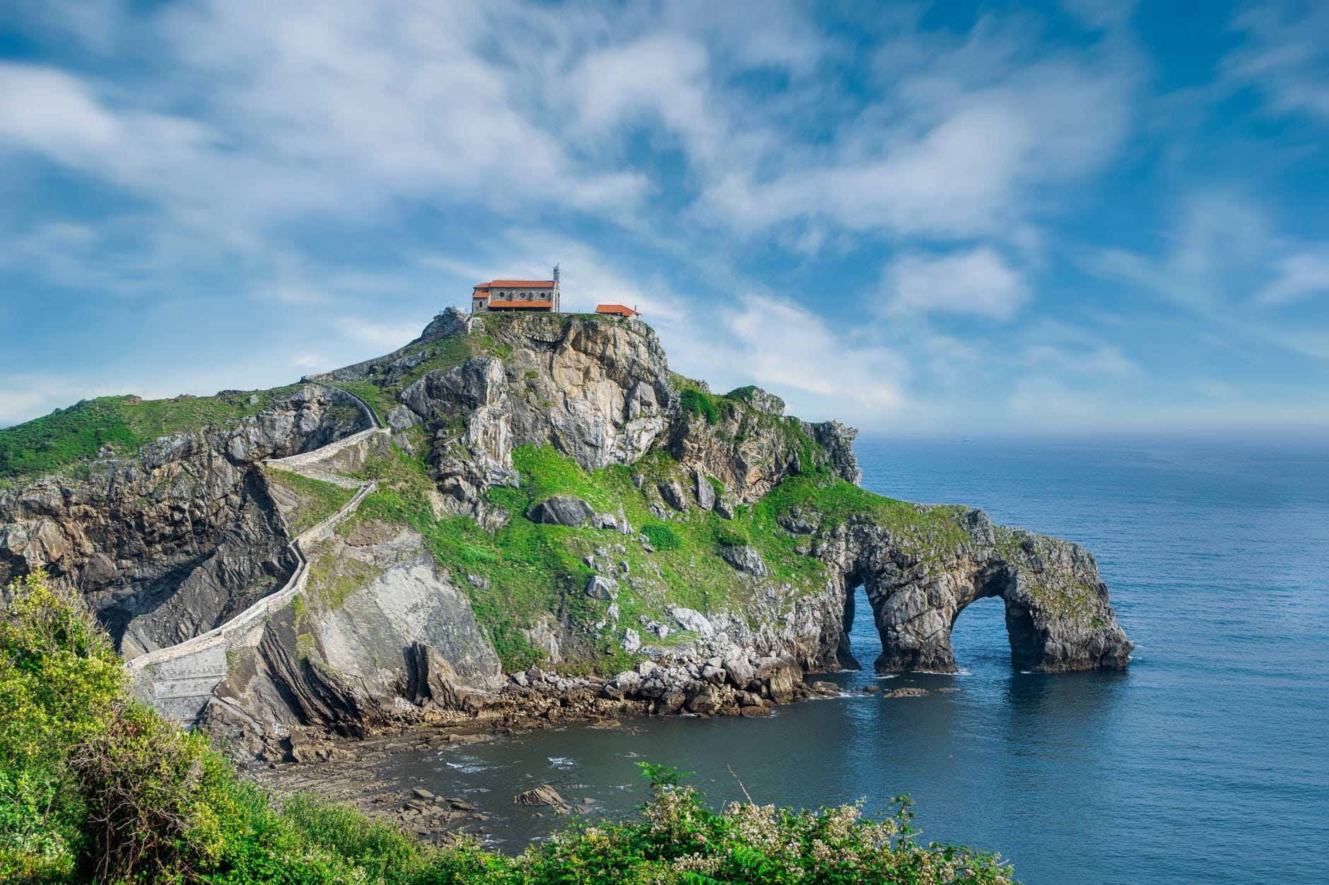 Die Einsiedelei San Juan de Gaztelugatxe thront auf einem dramatischen Felseiland, verbunden durch eine geschwungene Steinbrücke, umgeben vom blauen Meer unter strahlendem Himmel.