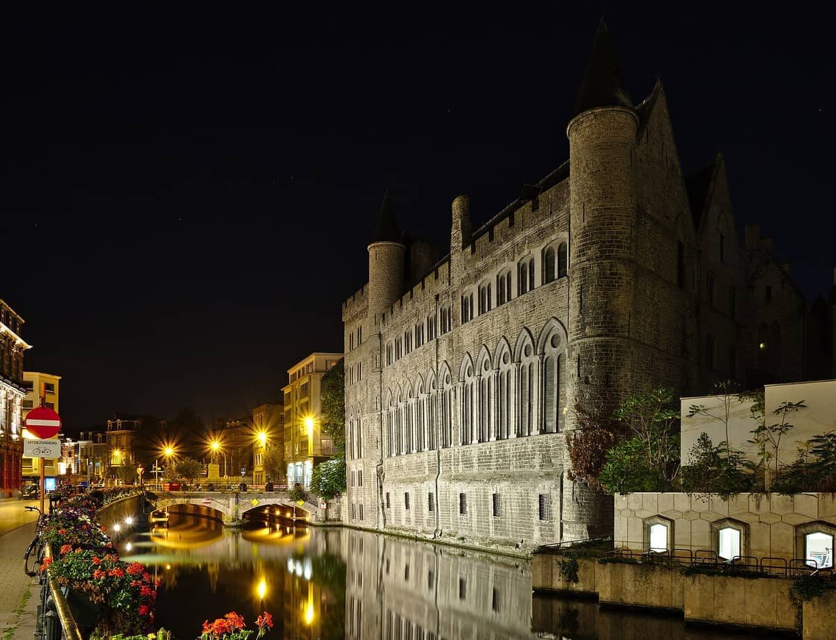 Geeraard de Duivelsteen’s stone facade and gothic towers reflected in the calm canal waters, illuminated by nighttime streetlights in Ghent.