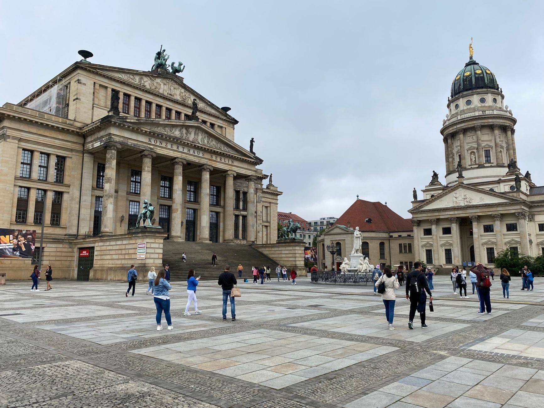Vista panoramica della piazza Gendarmenmarkt con persone che passeggiano, il Konzerthaus Berlin sulla sinistra e una cattedrale con cupola sulla destra sotto un cielo nuvoloso.