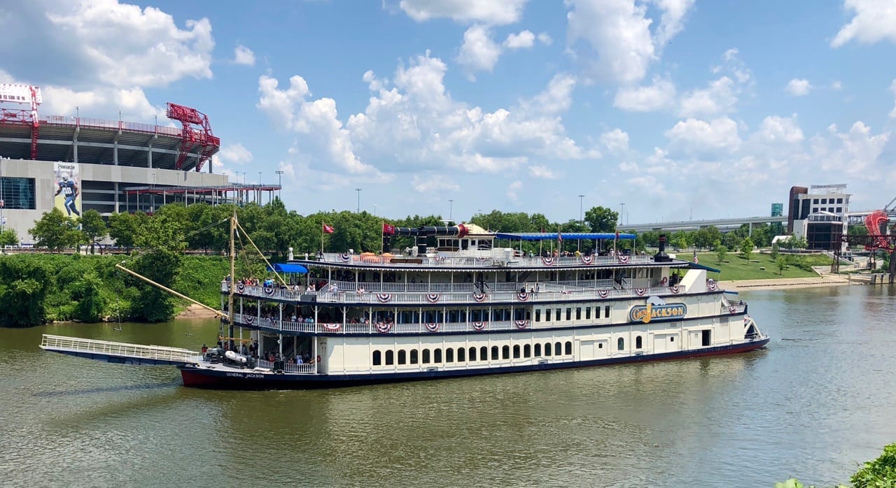 El barco espectáculo General Jackson, decorado con banderines americanos, navega por el río Cumberland bajo un cielo parcialmente nublado con un estadio al fondo.
