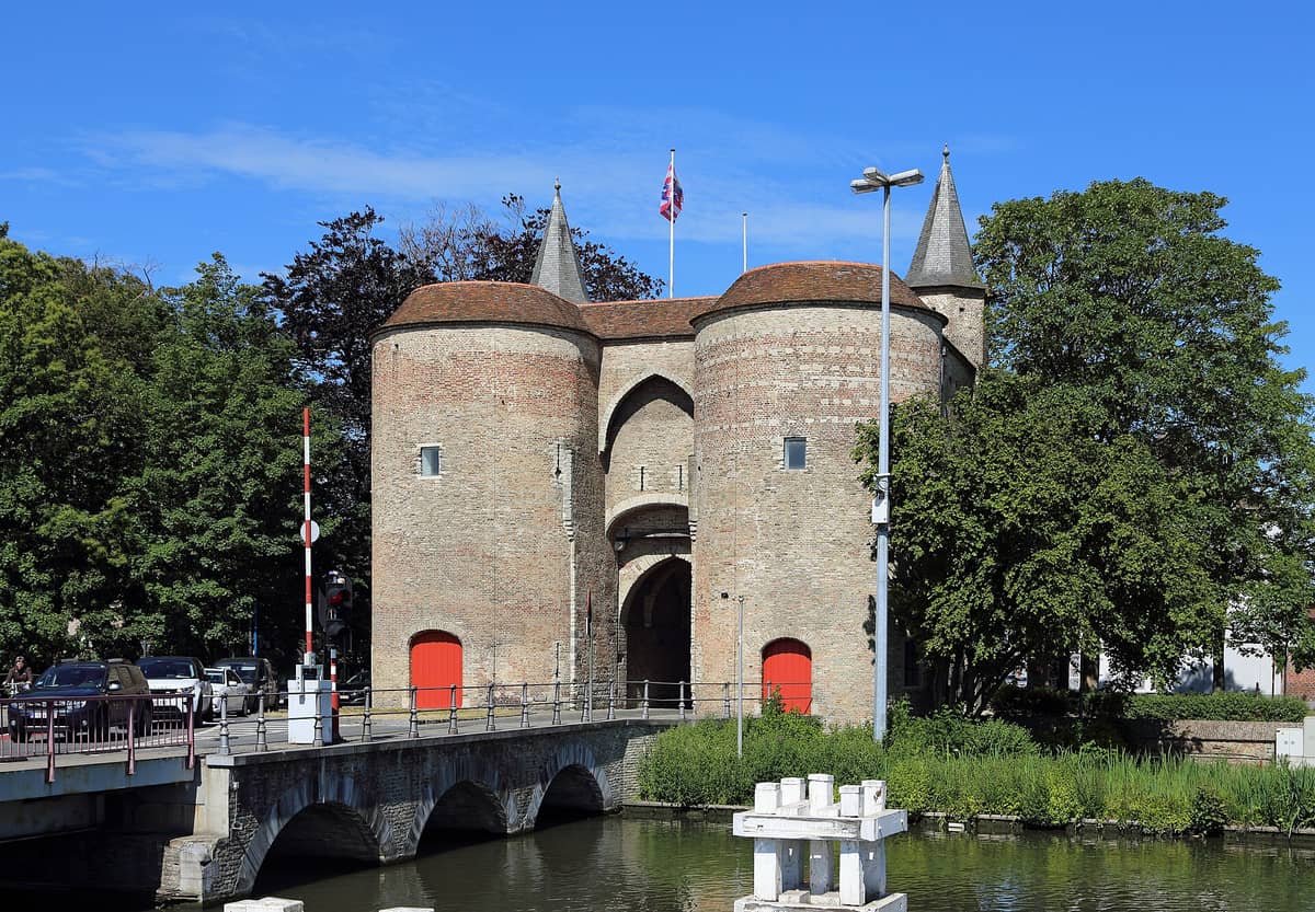 Front view of the Gentpoort (Ghent Gate) in Bruges, a medieval city gate with two round towers, red doors, a flag on top, and a canal bridge in front.