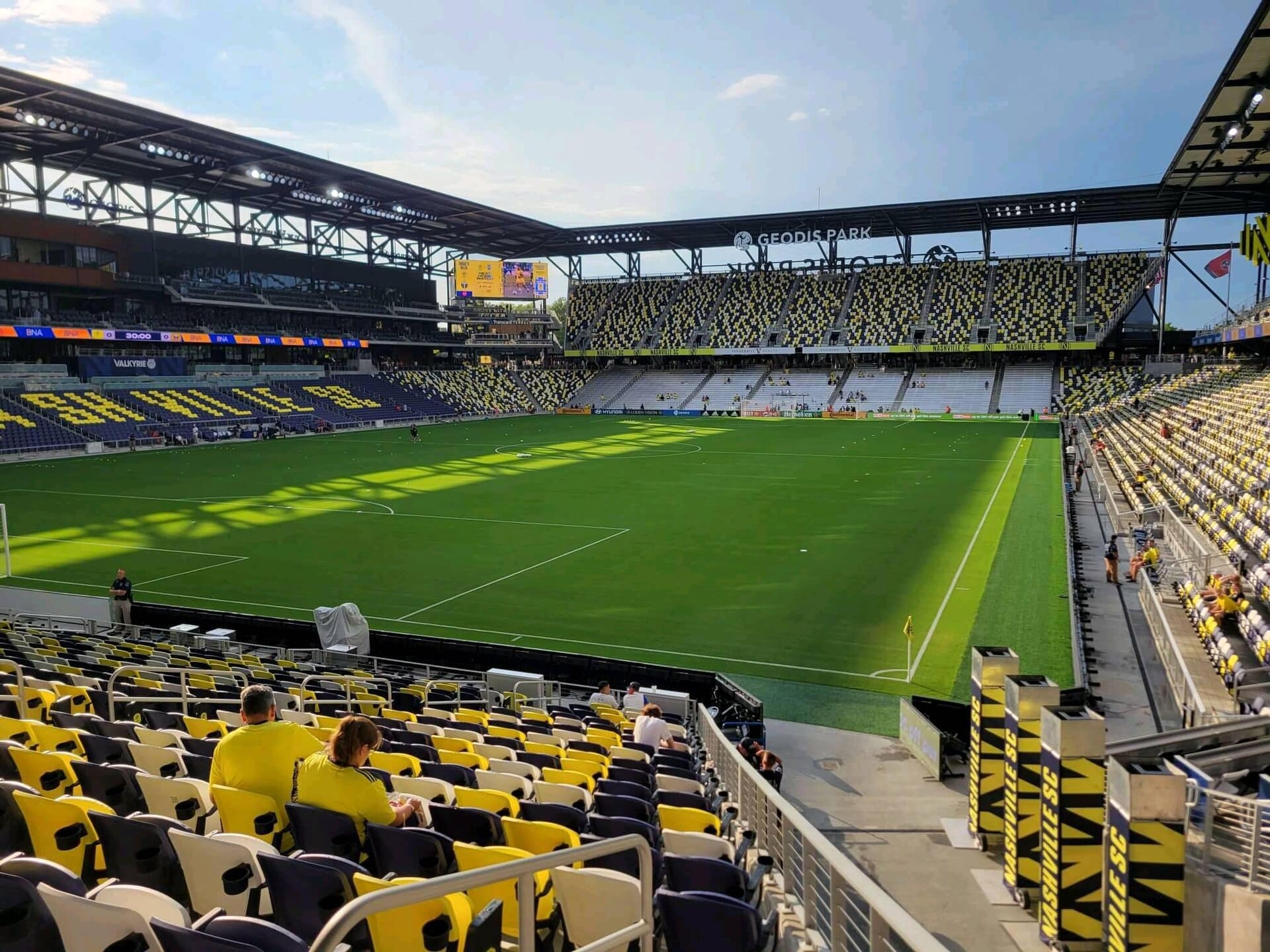 Vista amplia del estadio GEODIS Park en Nashville, con asientos amarillos y negros, campo de fútbol verde y algunos espectadores bajo el sol.
