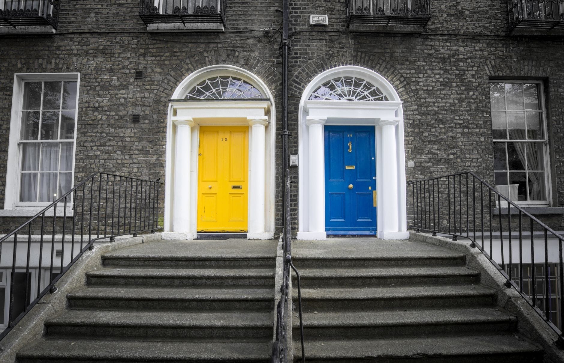 Duas entradas clássicas de casas georgianas em Dublin com portas amarelas e azuis vibrantes, bandeiras de vidro e tijolos à vista, destacando os traços arquitetônicos característicos do estilo georgiano.