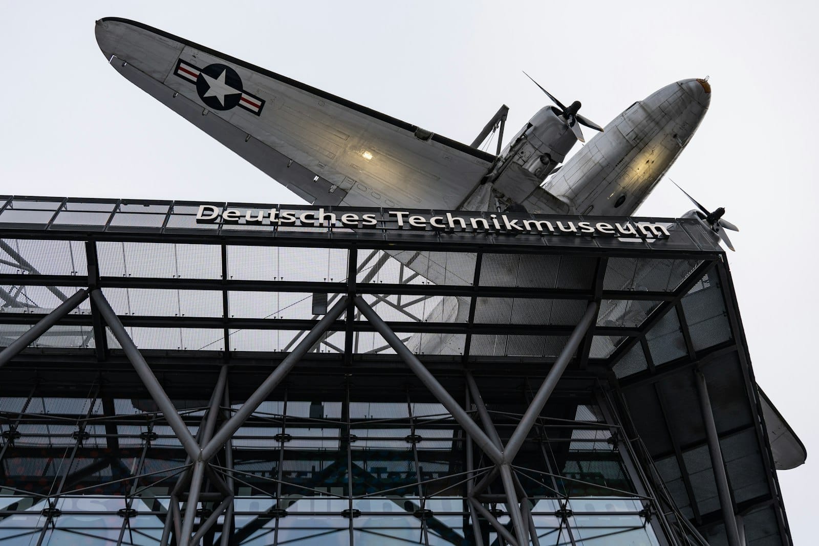 Esterno del Museo Tedesco della Tecnologia di Berlino con un aereo storico montato sopra l'ingresso e una struttura in acciaio visibile sotto.