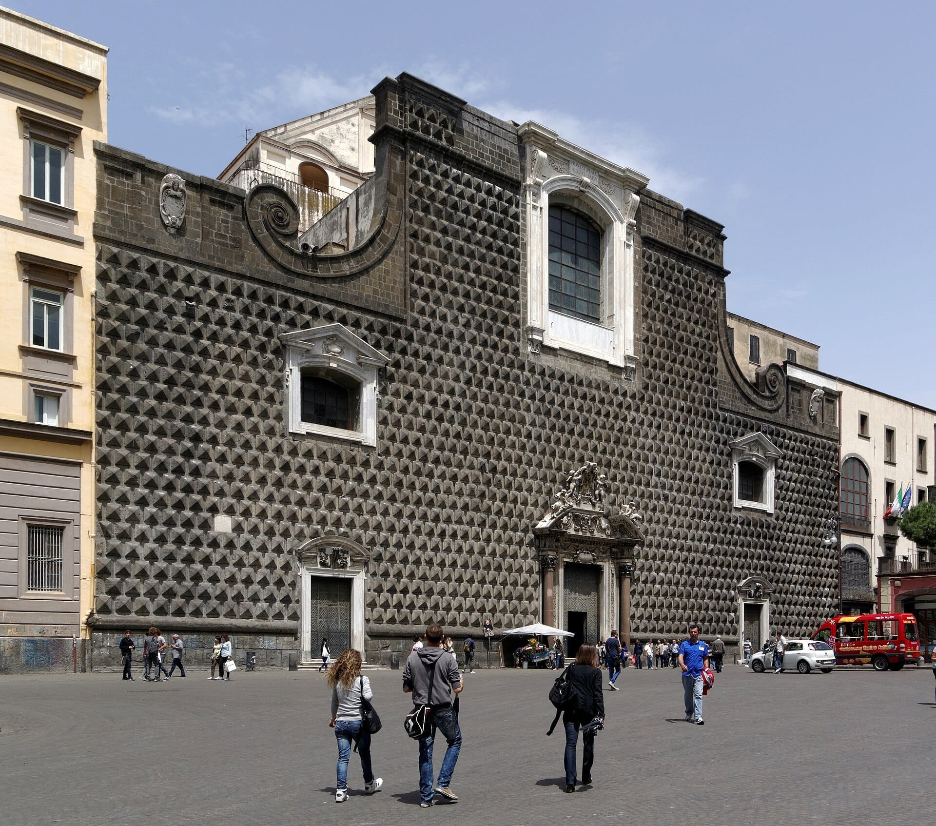 La Chiesa del Gesù Nuovo a Napoli con la sua distintiva facciata in bugnato a punta di diamante, incorniciata dagli edifici della città e dai visitatori che passeggiano nelle vicinanze in una giornata di sole.