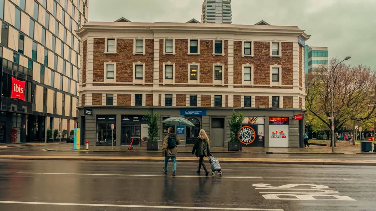 Menschen überqueren eine Stadtstraße im Adelaide CBD mit einem historischen Backsteingebäude und modernen Türmen im Hintergrund