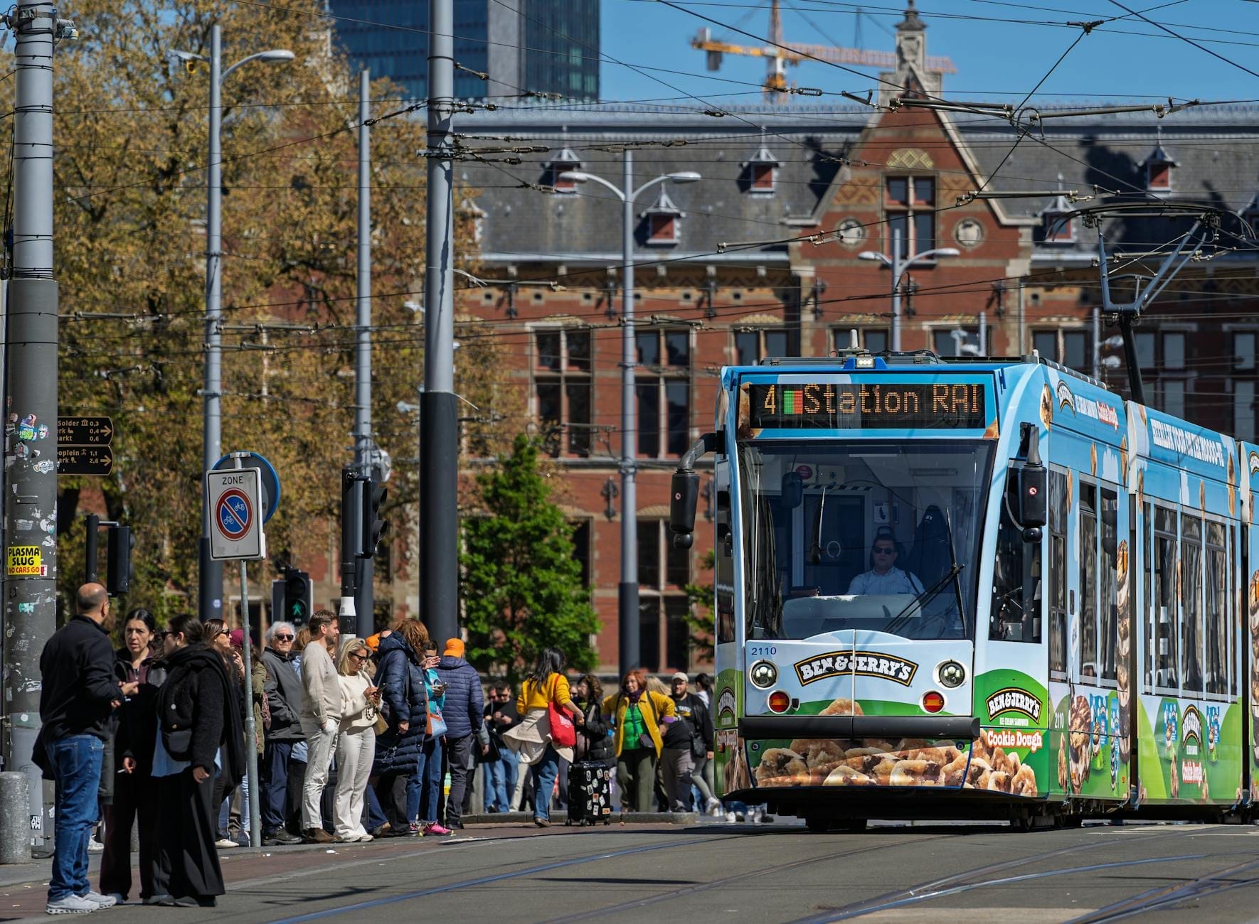 Tranvía de Amsterdam detenido en un concurrido andén con una gran multitud de personas esperando, frente a la arquitectura tradicional de la ciudad en un día soleado.