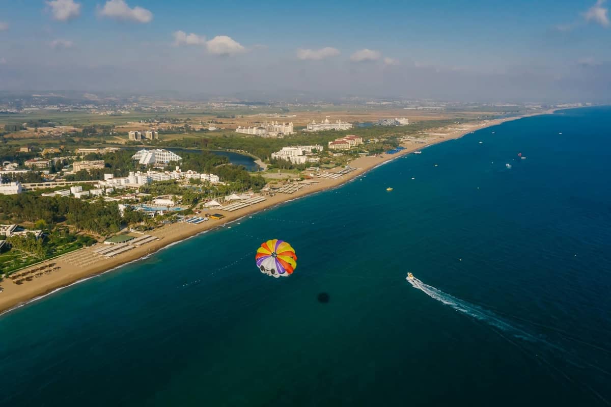 Aerial view of a Mediterranean coastal resort area in Antalya with hotels, beach, and a parasail over the blue sea.