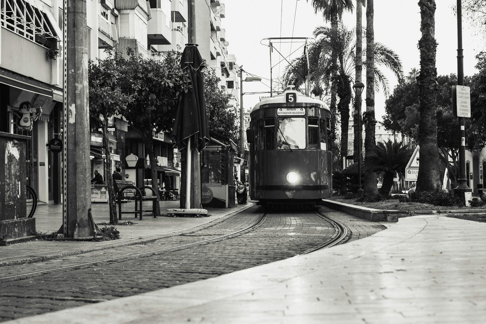 Foto em preto e branco de um bonde se aproximando no centro de Antalya, com palmeiras, prédios e pessoas nas calçadas.