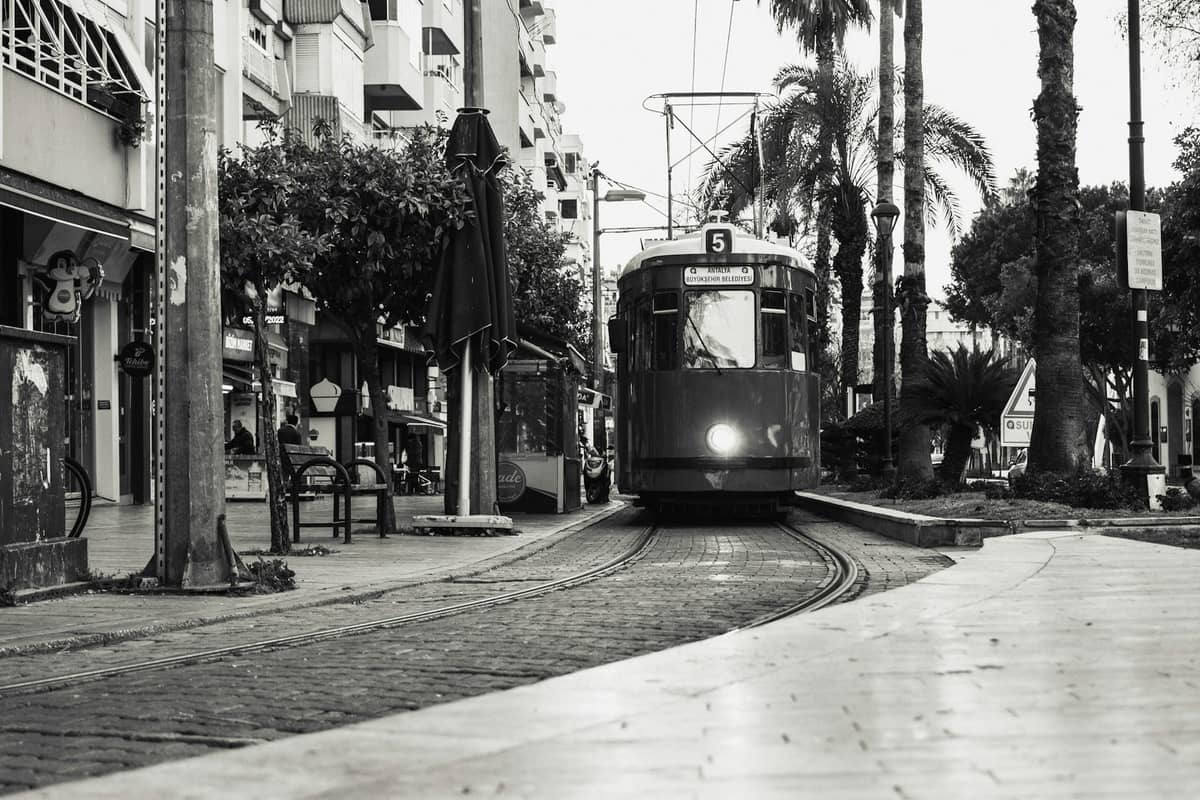 A vintage tram traveling down a palm-lined city street in Antalya, with pedestrians, buildings, and benches visible alongside.