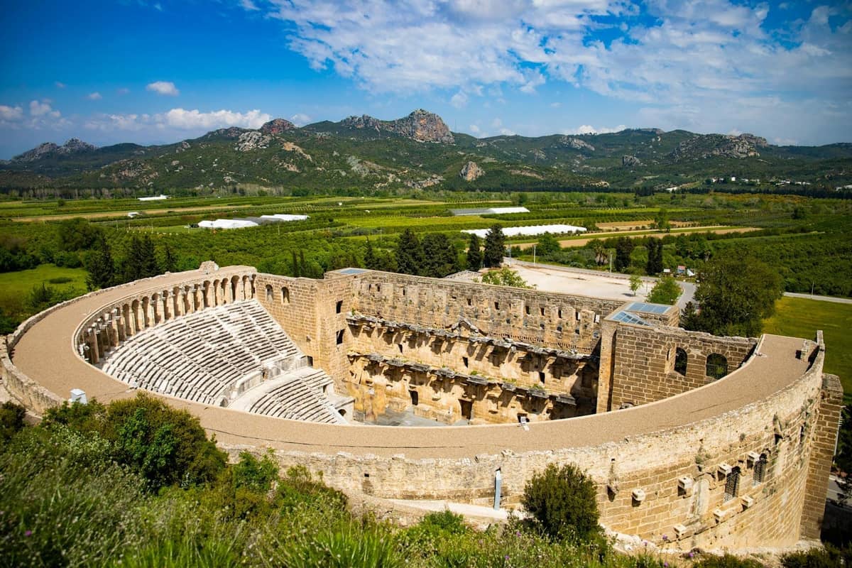 Wide view of the ancient Aspendos Theater near Antalya, surrounded by green countryside and distant mountains under a blue sky.