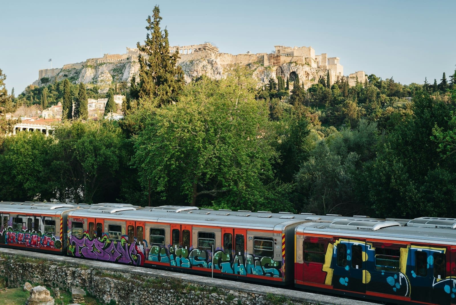 Um metrô colorido com grafite passa em frente a uma vegetação exuberante com a Acrópole ao fundo, simbolizando o transporte urbano e os pontos icônicos de Atenas.