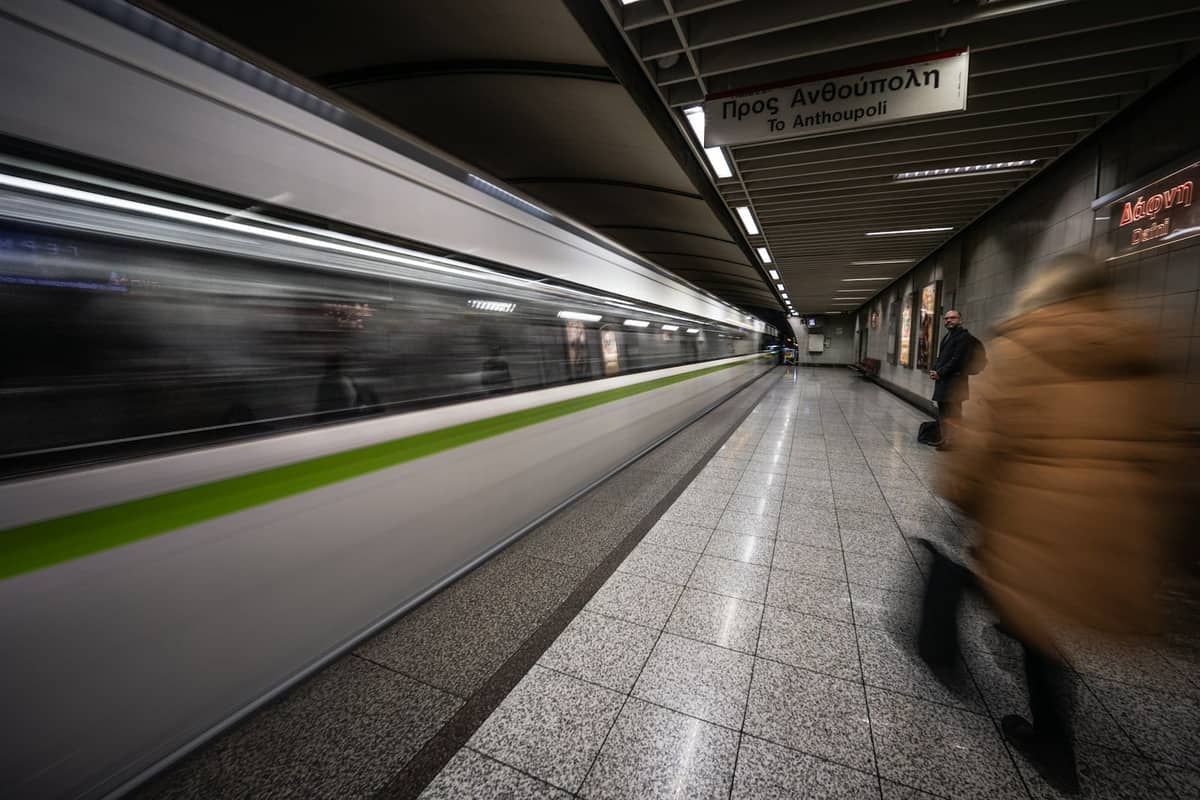 Innenansicht einer Athens Metro-Station mit einem grün gestreiften einfahrenden Zug und griechischen Schildern über einem gut beleuchteten Bahnsteig.