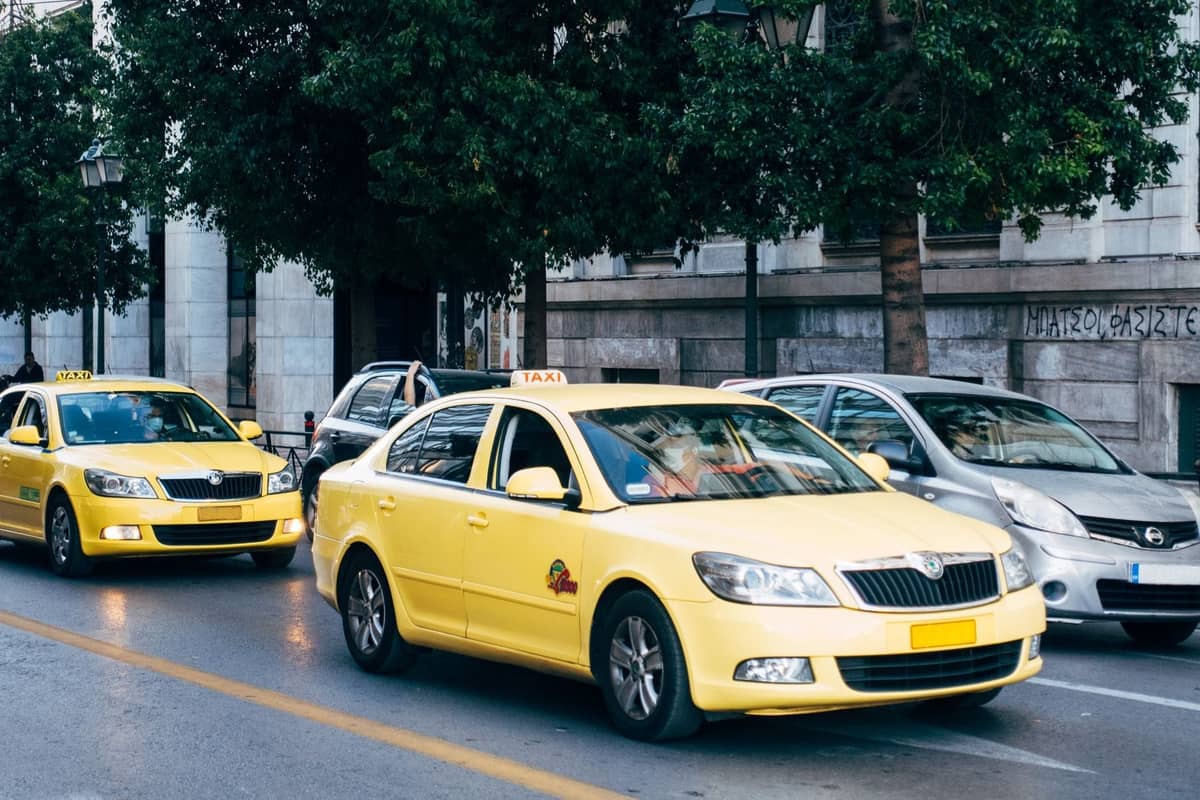 Gelbe Athener Taxis fahren auf einer Stadtstraße neben anderen Autos, vor einer Kulisse aus städtischen Gebäuden und Bäumen.