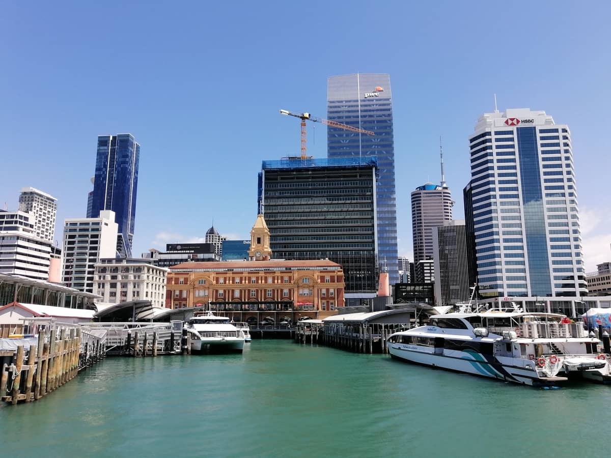 Terminal promowy w centrum Auckland z historycznym budynkiem promowym, nowoczesnymi wieżowcami i kilkoma promami zacumowanymi w żywych niebieskich wodach portu pod bezchmurnym niebem.
