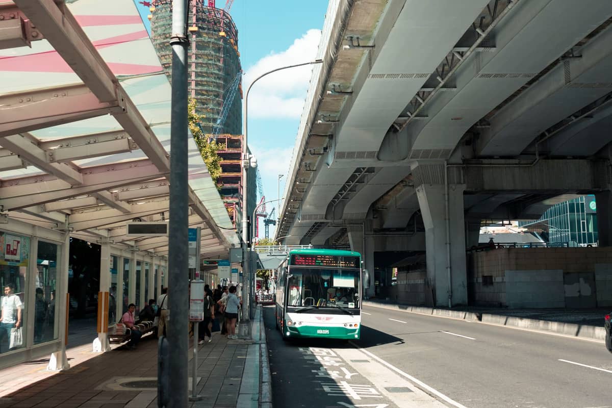 Autobus miejski przyjeżdża na zadaszony przystanek autobusowy w Auckland przy ruchliwej ulicy i estakadzie, gdzie oczekują pasażerowie.