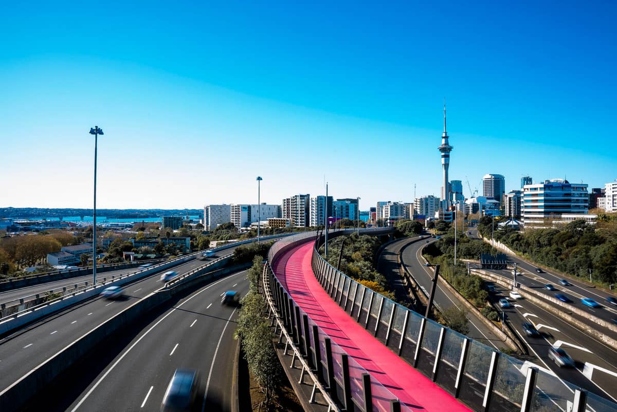 Blick auf die Skyline von Auckland bei Tag mit dem Sky Tower, Stadtgebäuden, einer belebten Autobahn und dem leuchtend pinken Lightpath-Radweg unter einem strahlend blauen Himmel.