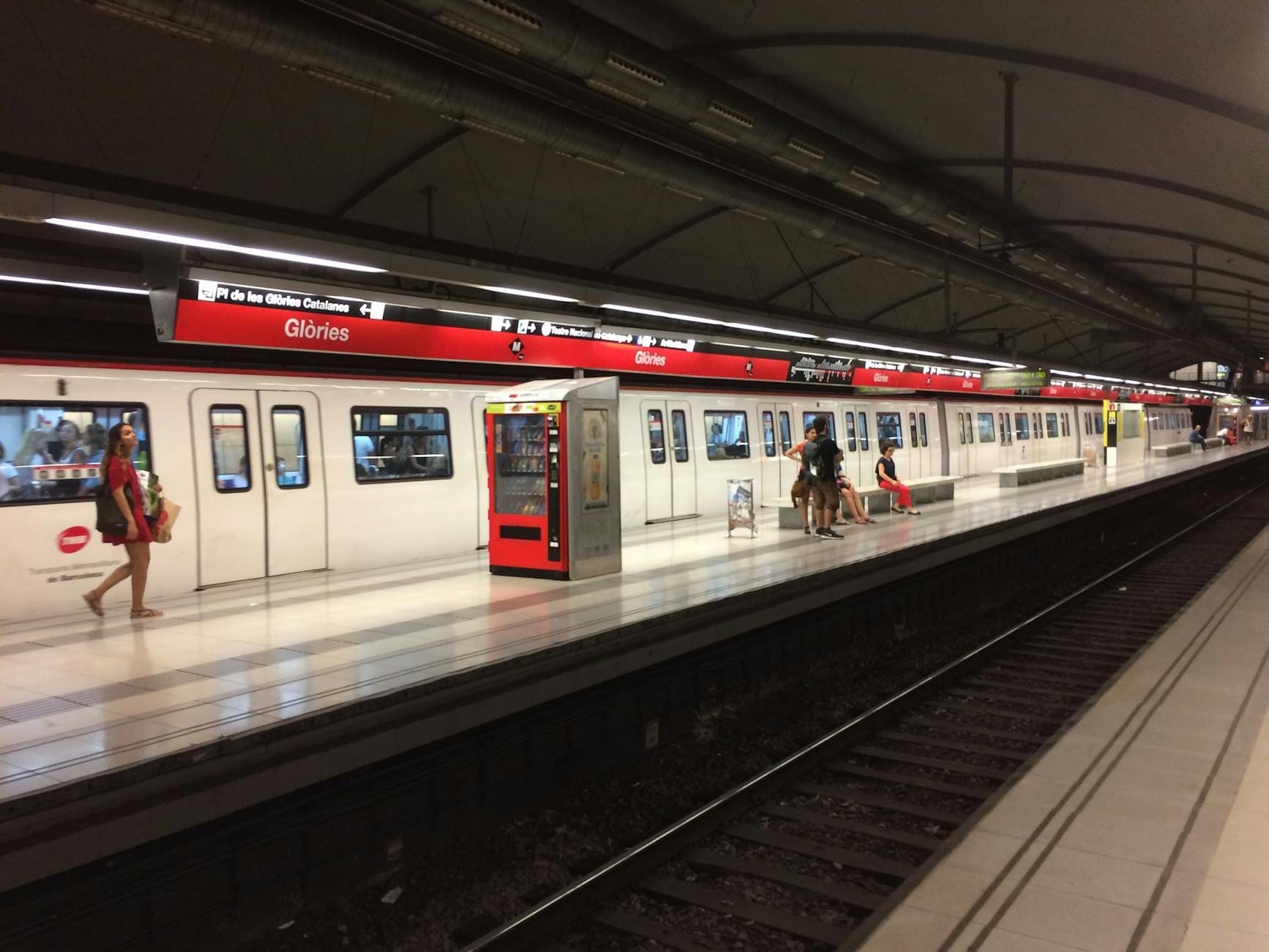 Wide view of Barcelona metro's Glòries station, with a train at the platform and passengers waiting or walking along the platform.