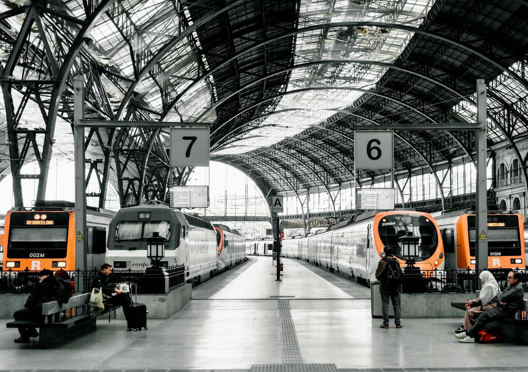 Wide view of Barcelona train station interior with multiple regional trains, large glass ceiling, passengers waiting and walking.