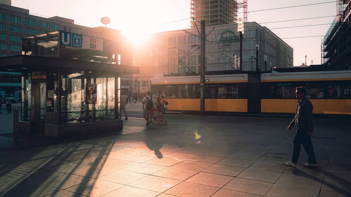 Berliner Straßenszene bei Sonnenuntergang mit einer gelben Straßenbahn, U-Bahn-Eingang, Fußgängern und Stadtgebäuden im Hintergrund.