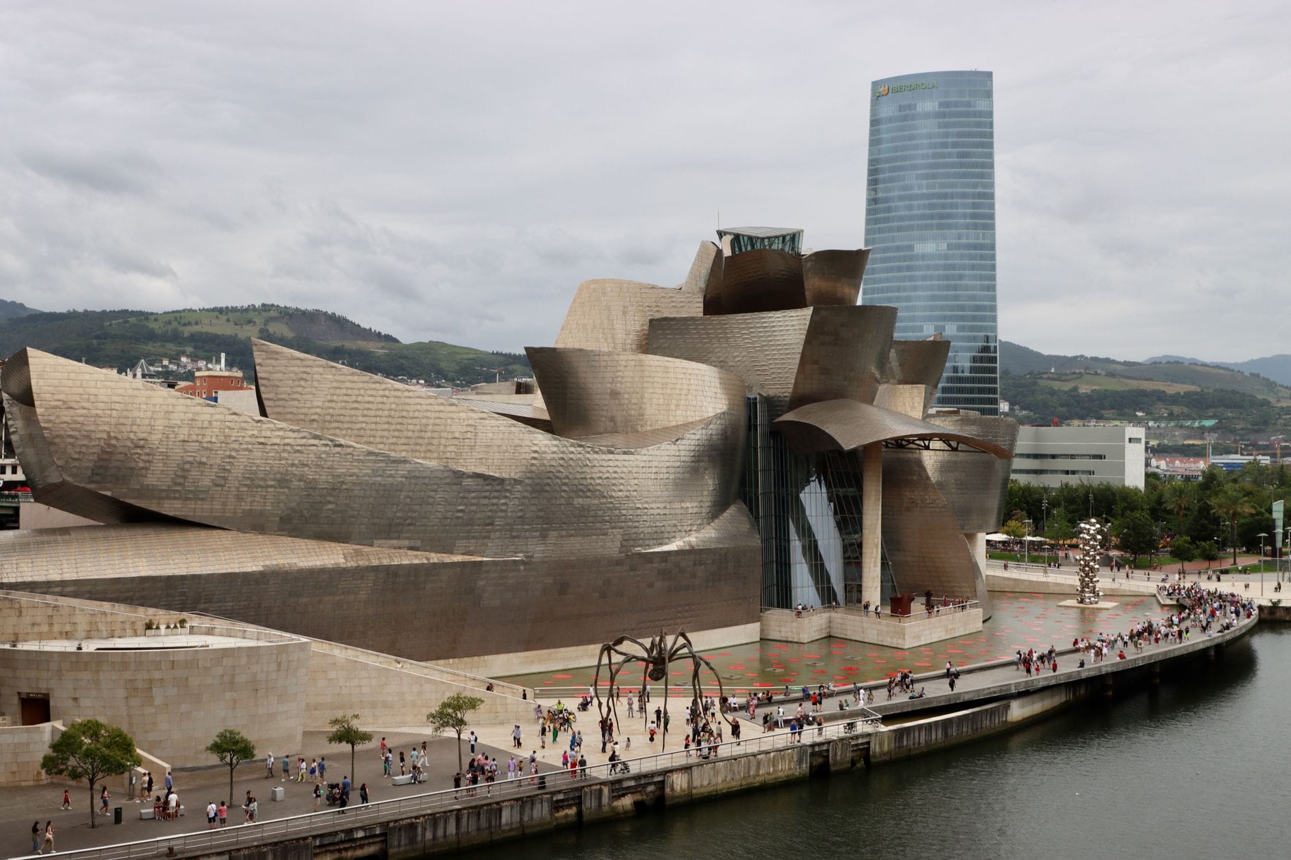 Breite Uferpromenade neben dem Guggenheim Museum Bilbao mit Spaziergängern und dem Torre Iberdrola im Hintergrund.