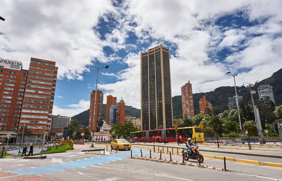 Bogotá city street with a red TransMilenio bus, taxi, motorbike, skyscrapers, and mountains under a cloudy sky.