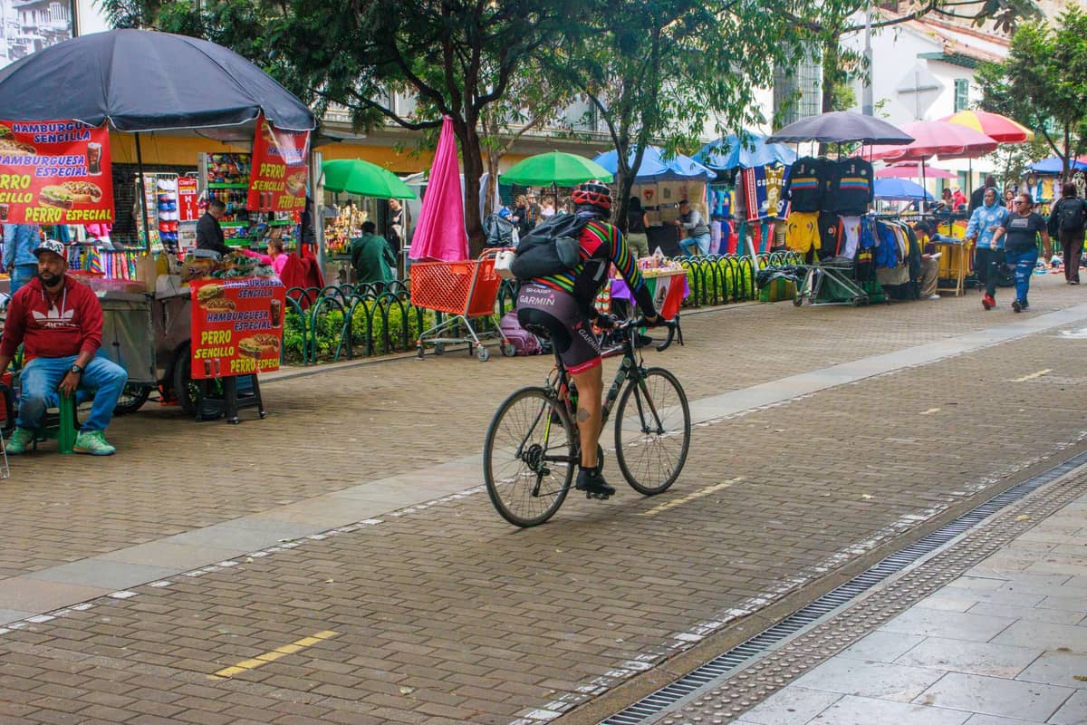 Cyclist riding on a bike lane through a lively Bogotá street lined with colorful street vendors and market stalls.