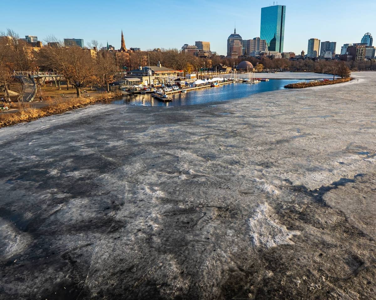 Zugefrorener Charles River mit Anlegestellen und der Skyline von Boston im Hintergrund, mit Teilen des Radwegs am Flussufer.