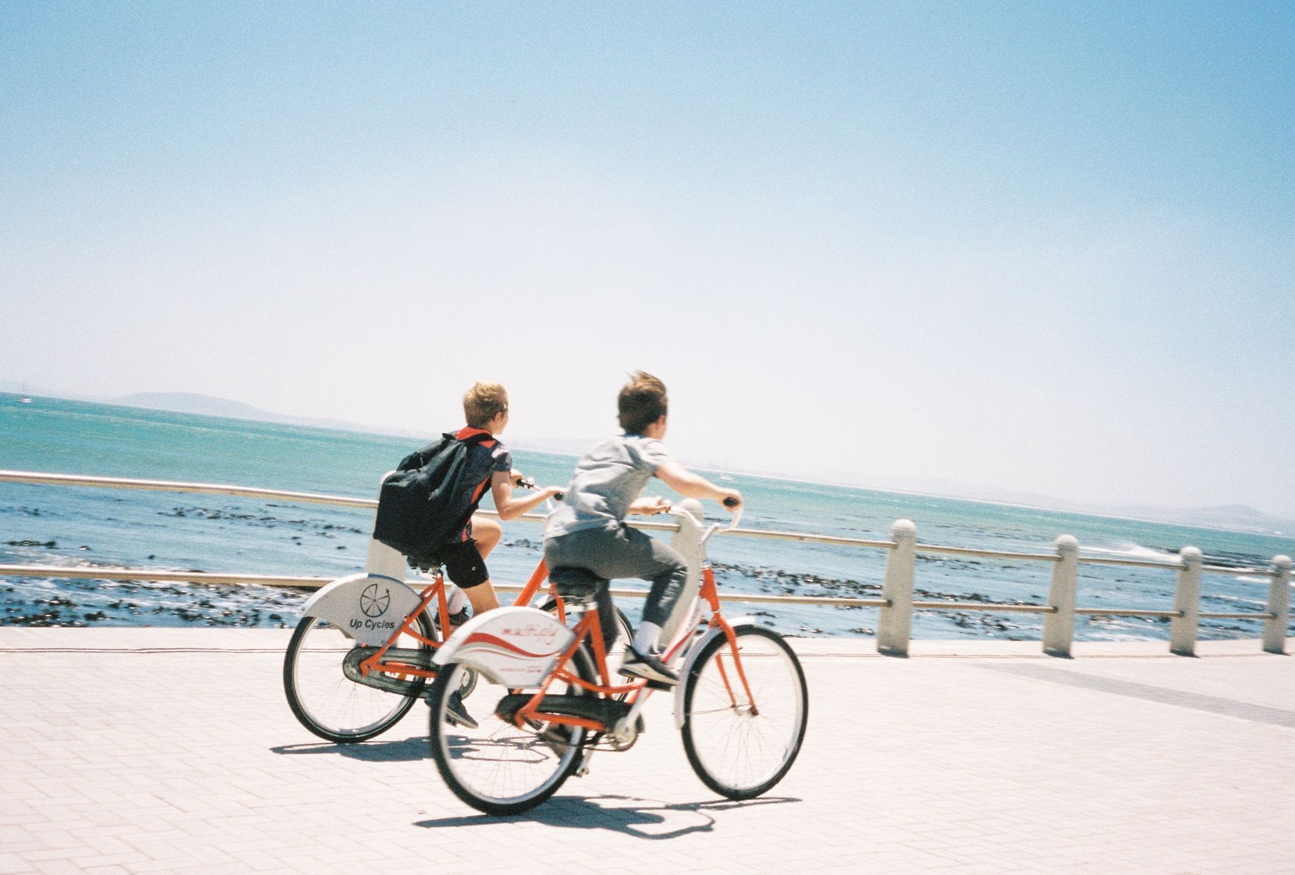 Zwei Personen fahren auf Mietfahrrädern entlang einer gepflasterten Promenade am Meer, mit strahlend blauem Himmel und Geländer, was an Küstenradfahren erinnert.