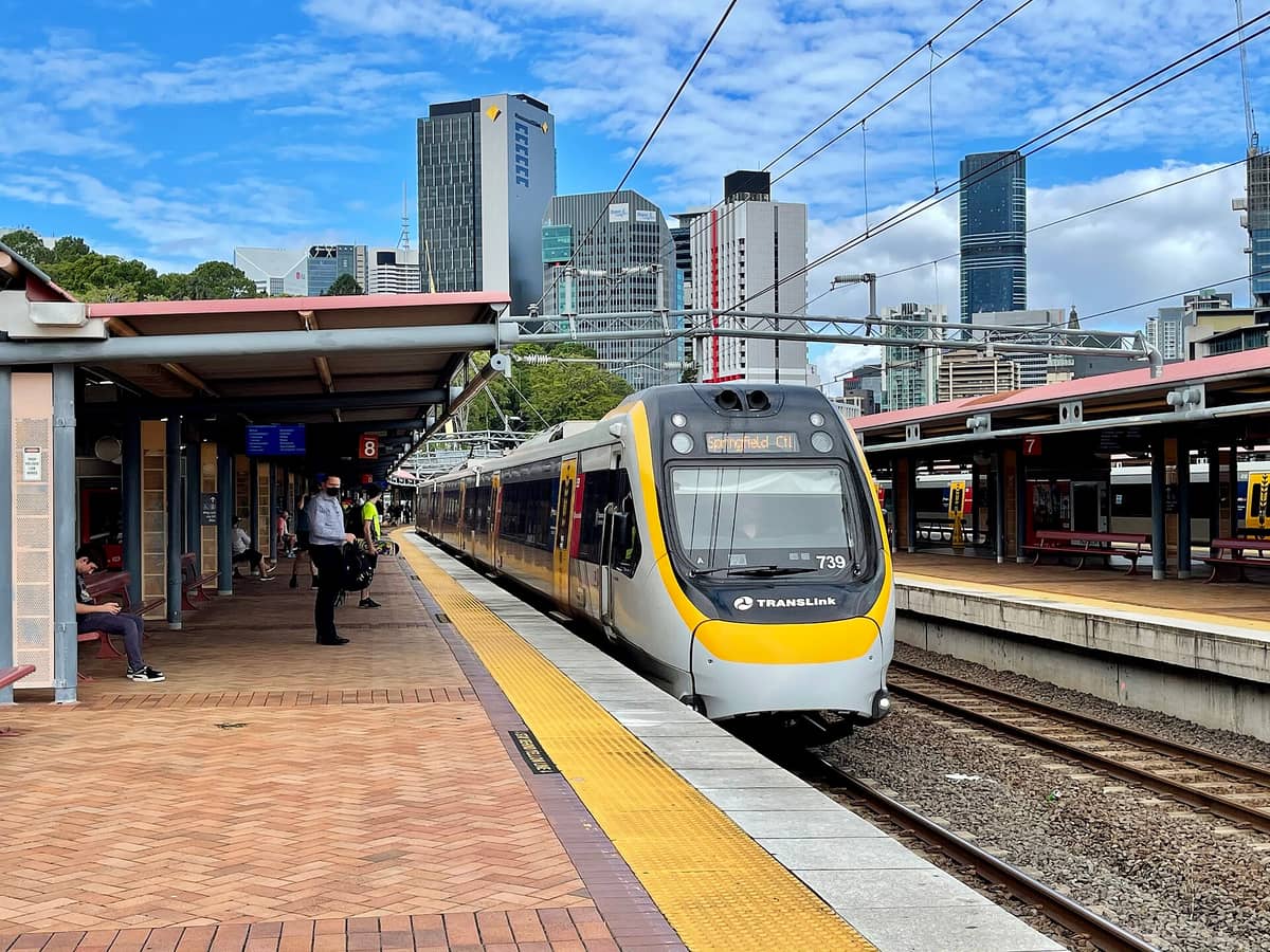 Modern Translink commuter train at a Brisbane city station with passengers waiting and city skyscrapers in the background.