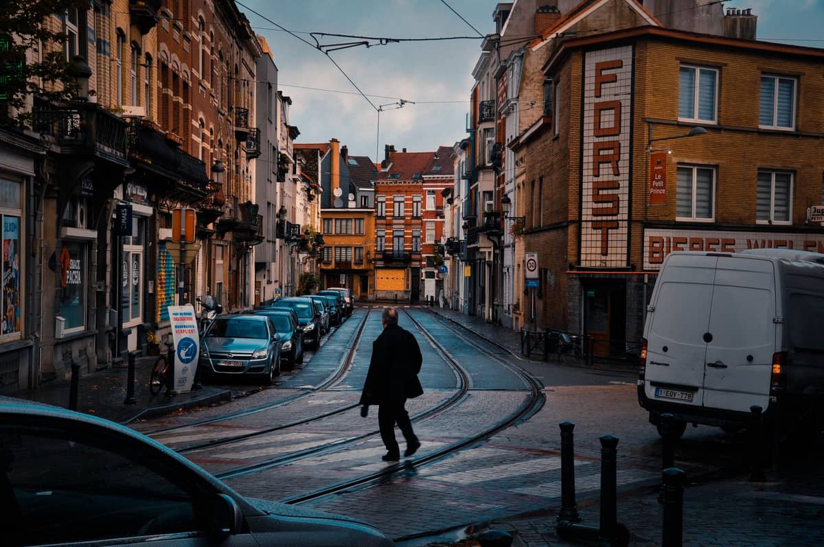Uma rua de Bruxelas com trilhos de bonde, fios elétricos aéreos, carros estacionados, uma faixa de pedestres e prédios clássicos da cidade, representando a infraestrutura de transporte urbano.
