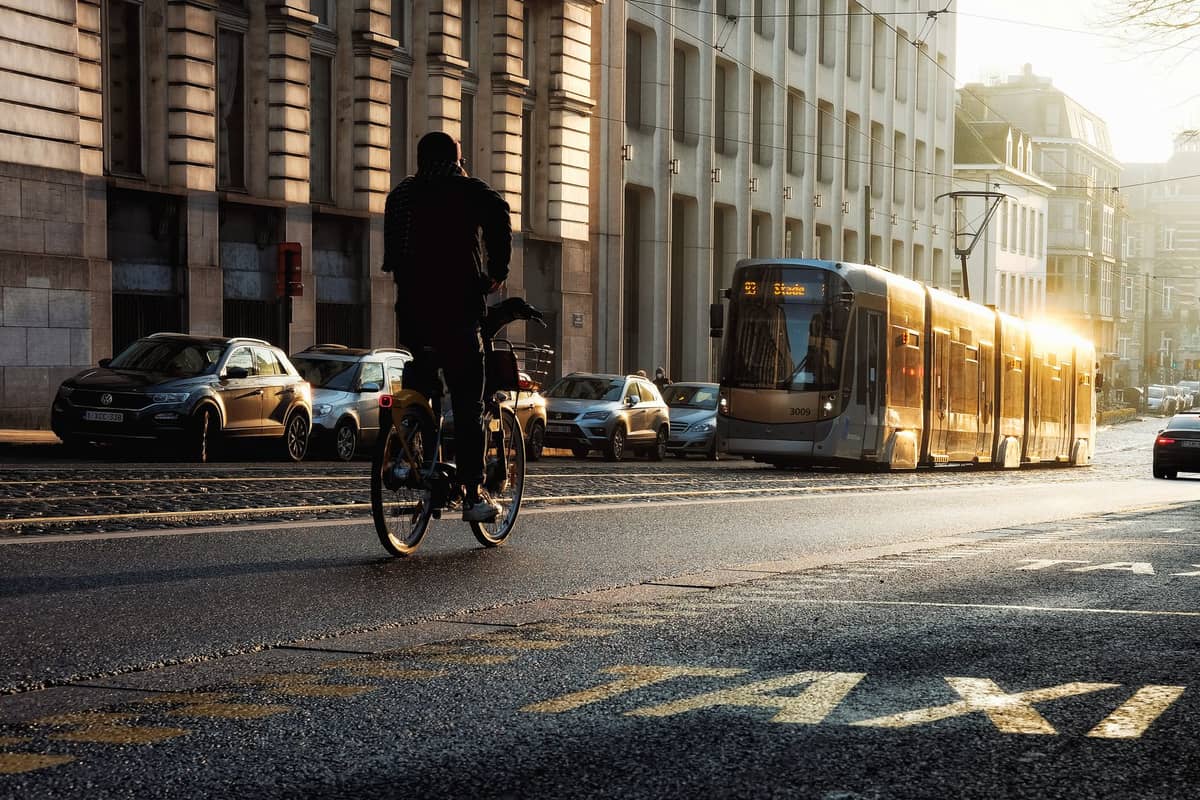 Um moderno elétrico de Bruxelas ao sol passando por uma rua da cidade com ciclista, carros e edifícios urbanos visíveis.