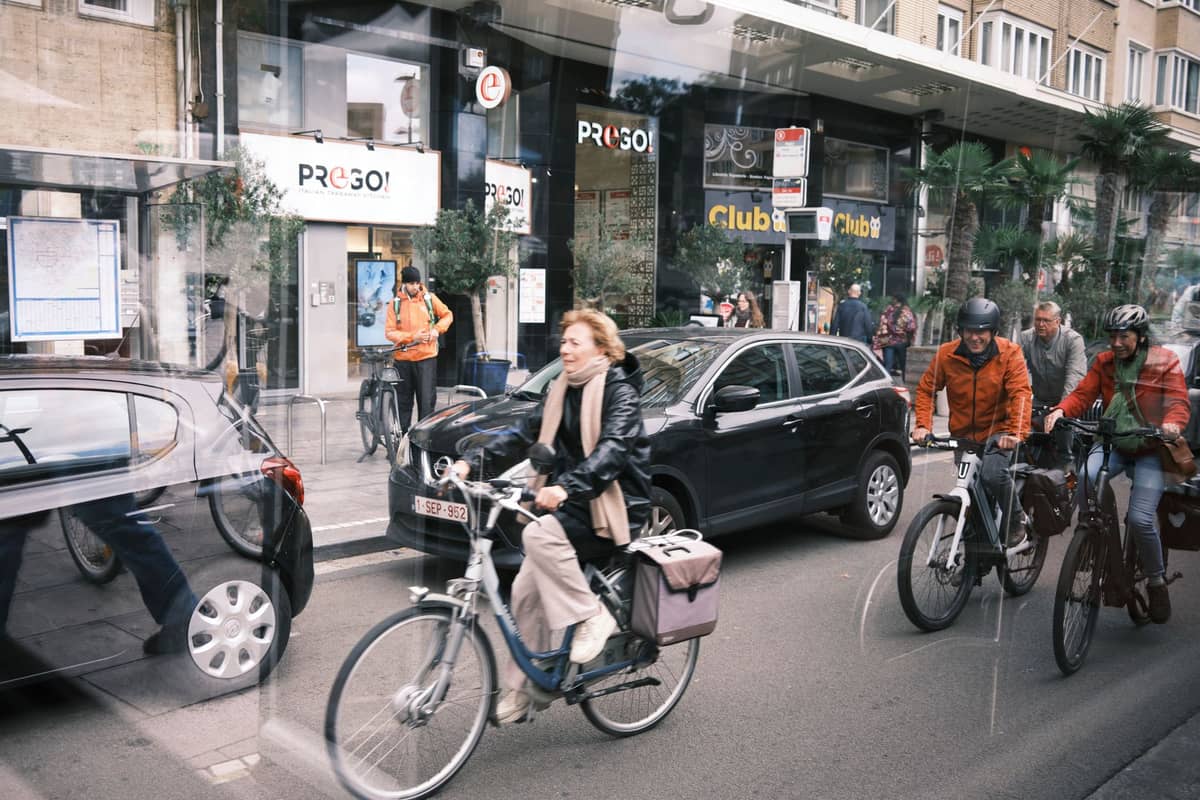 Vários ciclistas pedalam por uma rua de Bruxelas com edifícios, carros estacionados e pedestres visíveis ao fundo.