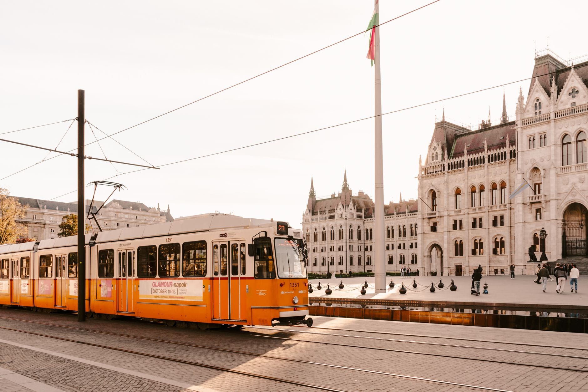 Leuchtend gelbe Straßenbahn fährt an einem sonnigen Tag vor dem Budapester Parlamentsgebäude vorbei, mit klar sichtbaren Stadtdetails und Gleisen.