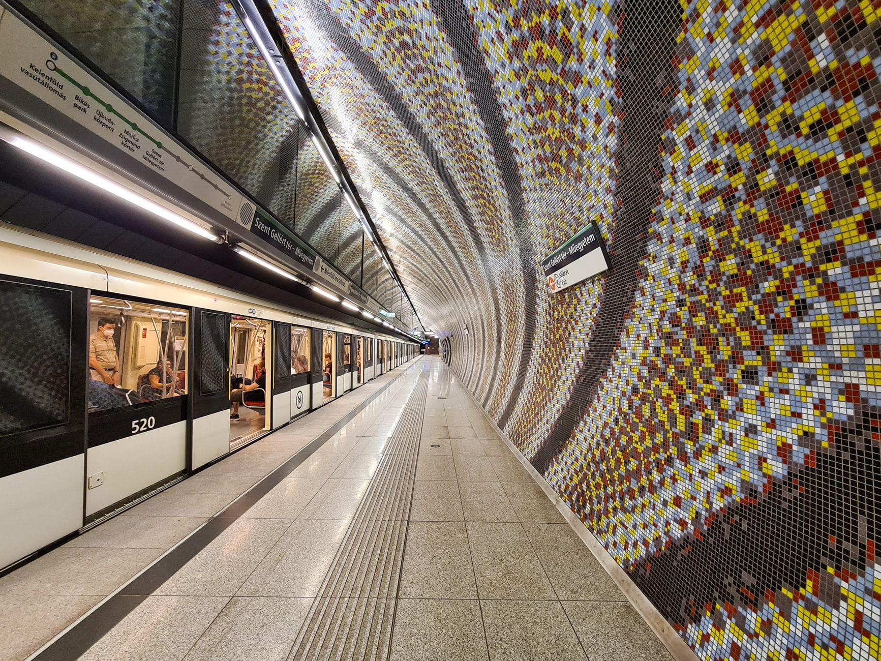 Hell erleuchtete Budapest-U-Bahnstation mit Mosaikwänden, Bahnsteig und einem am Bahnsteig haltenden U-Bahn-Zug.