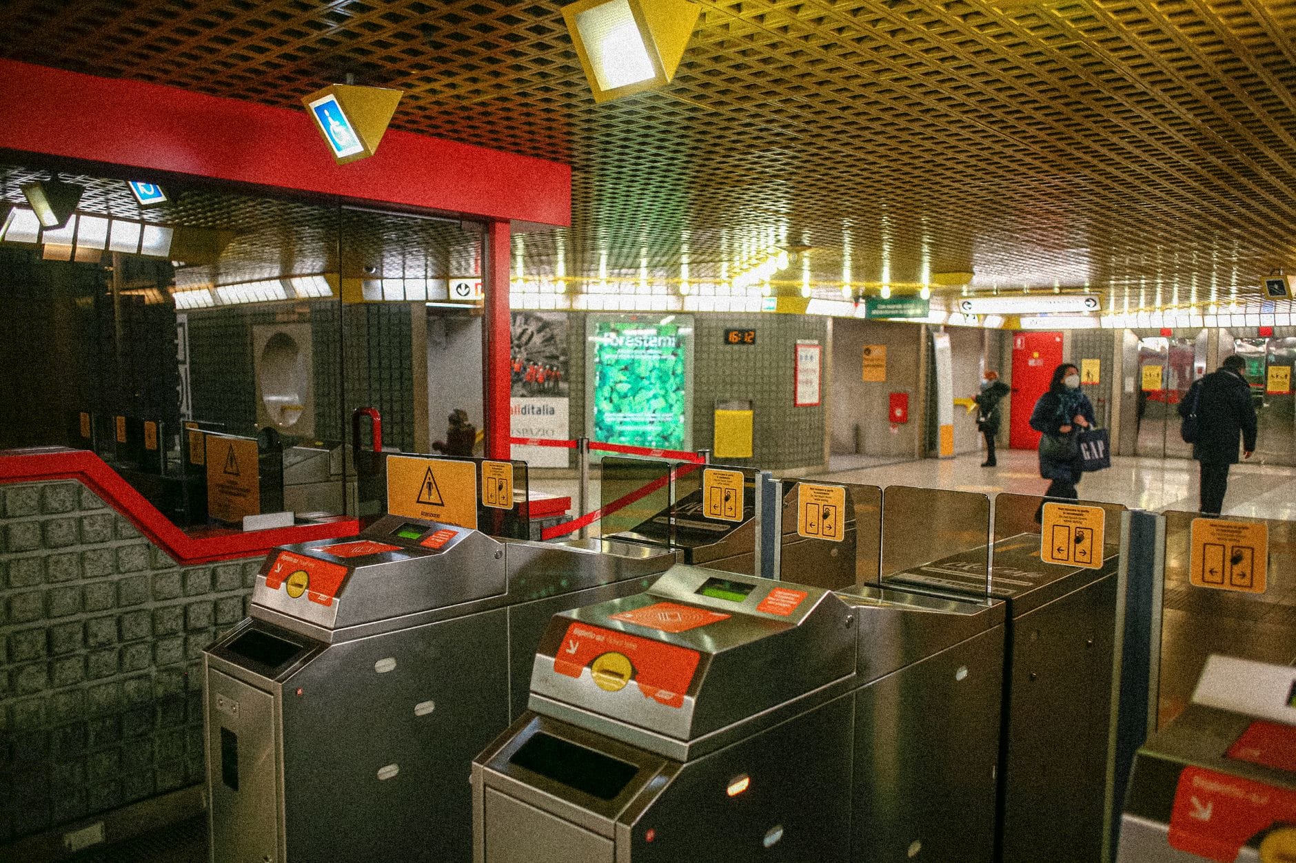 Innenansicht einer U-Bahn-Station in Budapest mit orangefarbenen Fahrsperren, eintretenden Personen und bunter moderner Beschilderung darüber
