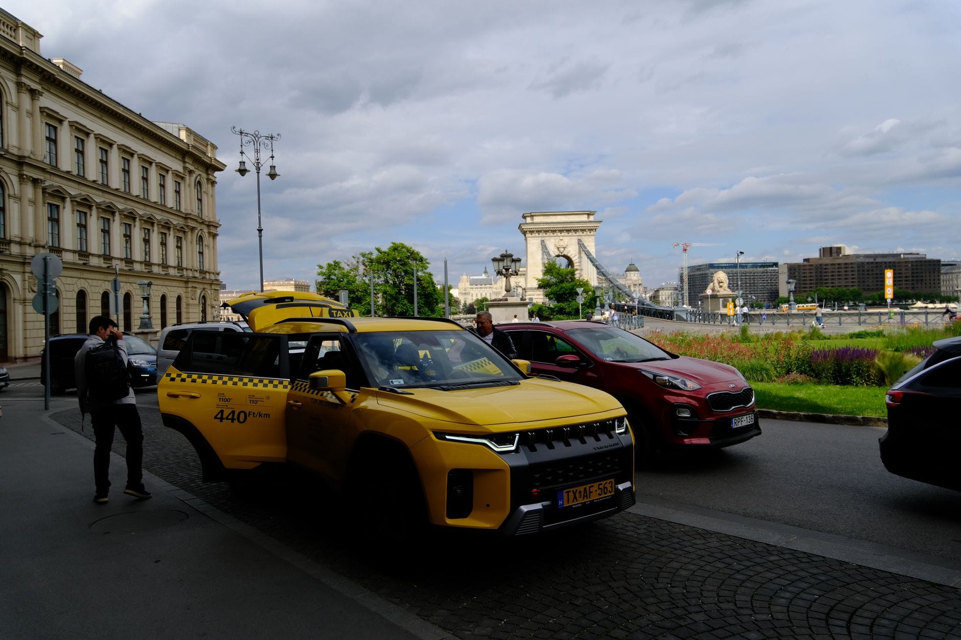 Ein gelbes Budapester Taxi neben der Kettenbrücke mit Stadtsehenswürdigkeiten und der Donau an einem bewölkten Tag.