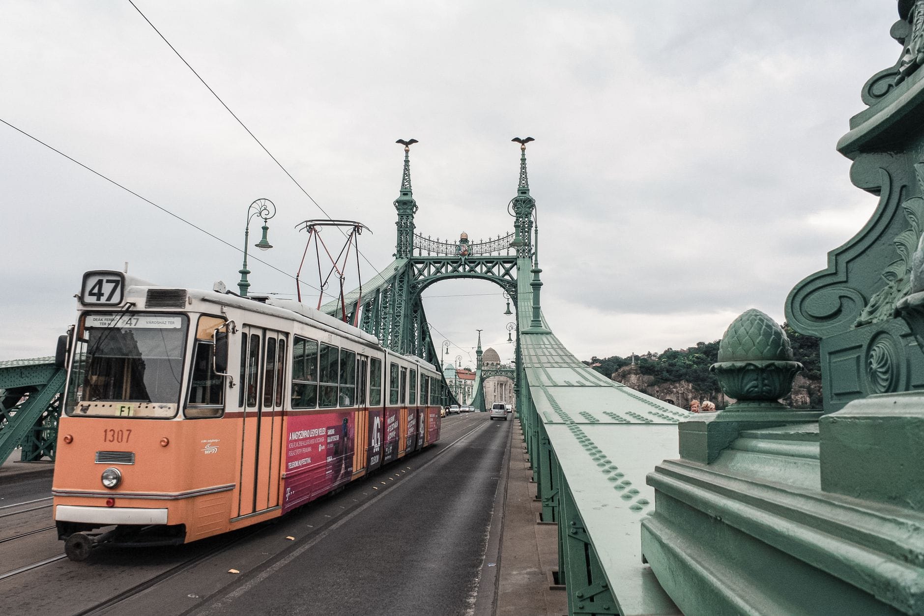 Eine klassische Budapester Straßenbahn überquert die Freiheitsbrücke mit ihren verzierten grünen Geländern, markanter Architektur und Stadtpanorama im Hintergrund an einem bewölkten Tag.