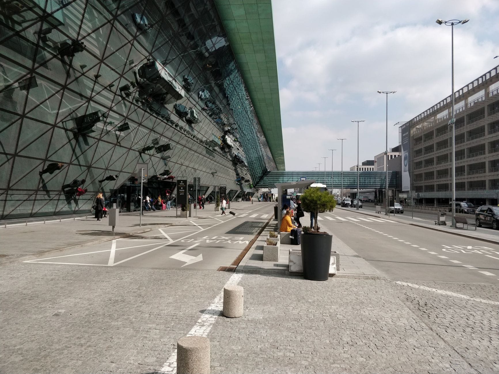Außenansicht des Terminals des Flughafens Budapest mit Glasfassade, Menschen, Fahrspuren und Gehweg unter bewölktem Himmel