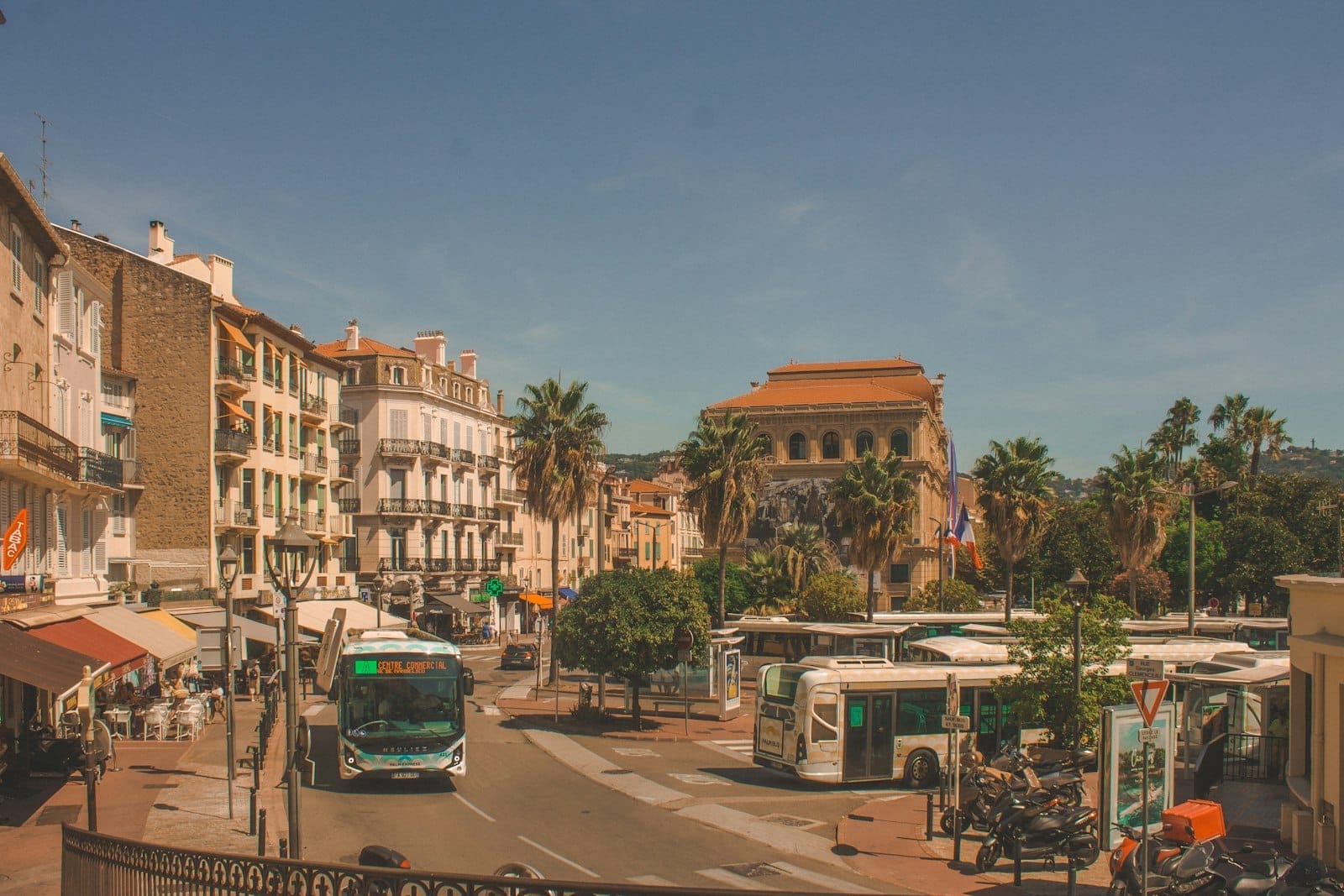 Rua movimentada de Cannes com ônibus, um coletivo verde, palmeiras e pedestres, mostrando as opções de transporte urbano sob um céu azul.
