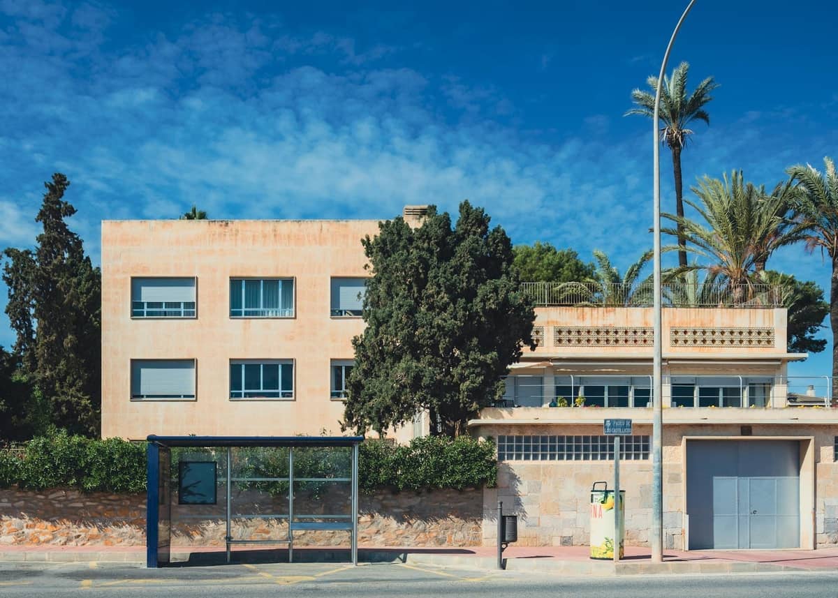 Eine gläserne Bushaltestelle auf einer sonnigen Straße vor Gebäuden im mediterranen Stil und Palmen.