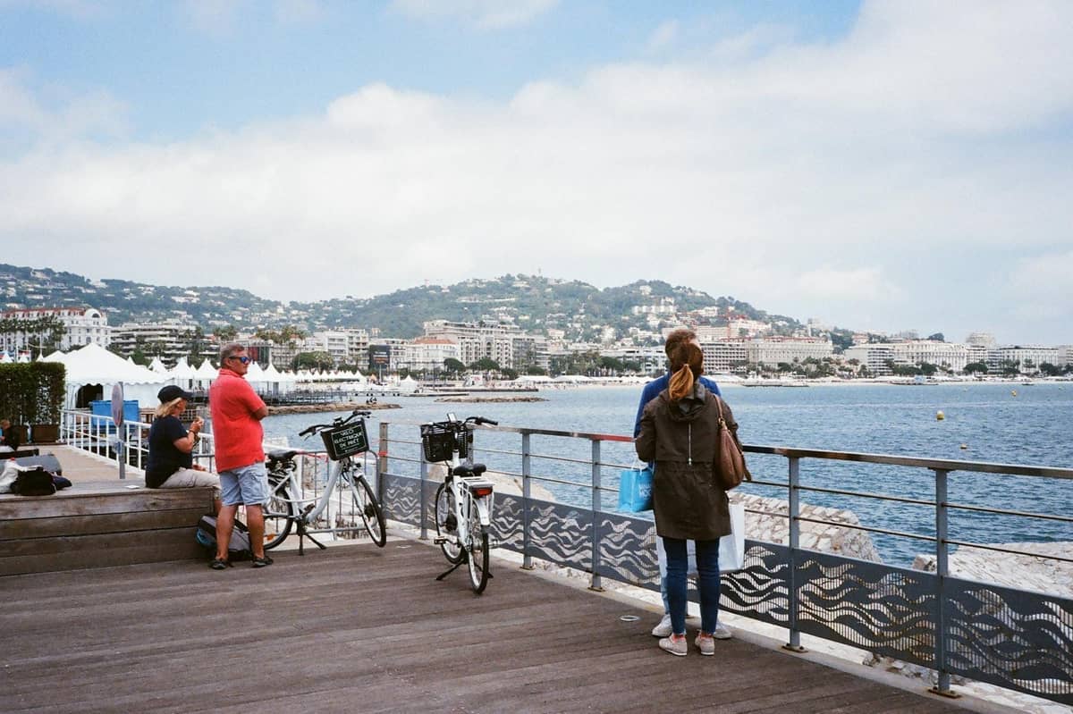 Menschen mit Leihfahrrädern auf einem Strandweg am Meer in Cannes, mit der Stadt und den Hügeln im Hintergrund.