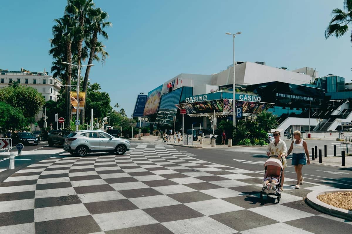 Menschen, die auf einer breiten Straße nahe dem Palais des Festivals in Cannes mit Palmen und einem auffälligen Zebrastreifen spazieren.